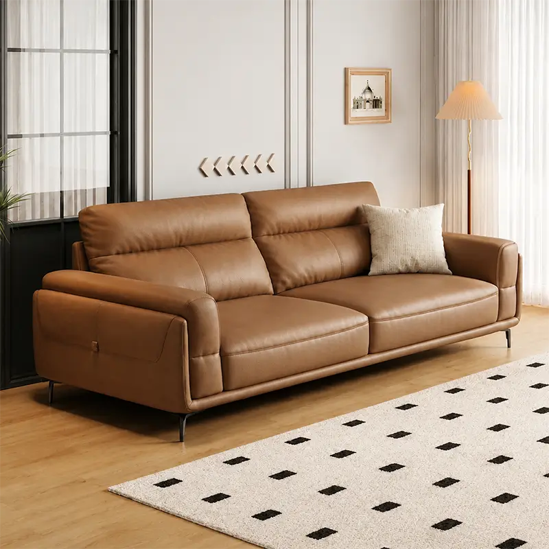 A modern three-seat sofa upholstered in warm tan leather, styled in a contemporary living room with a cream throw pillow, a patterned rug, and a floor lamp.